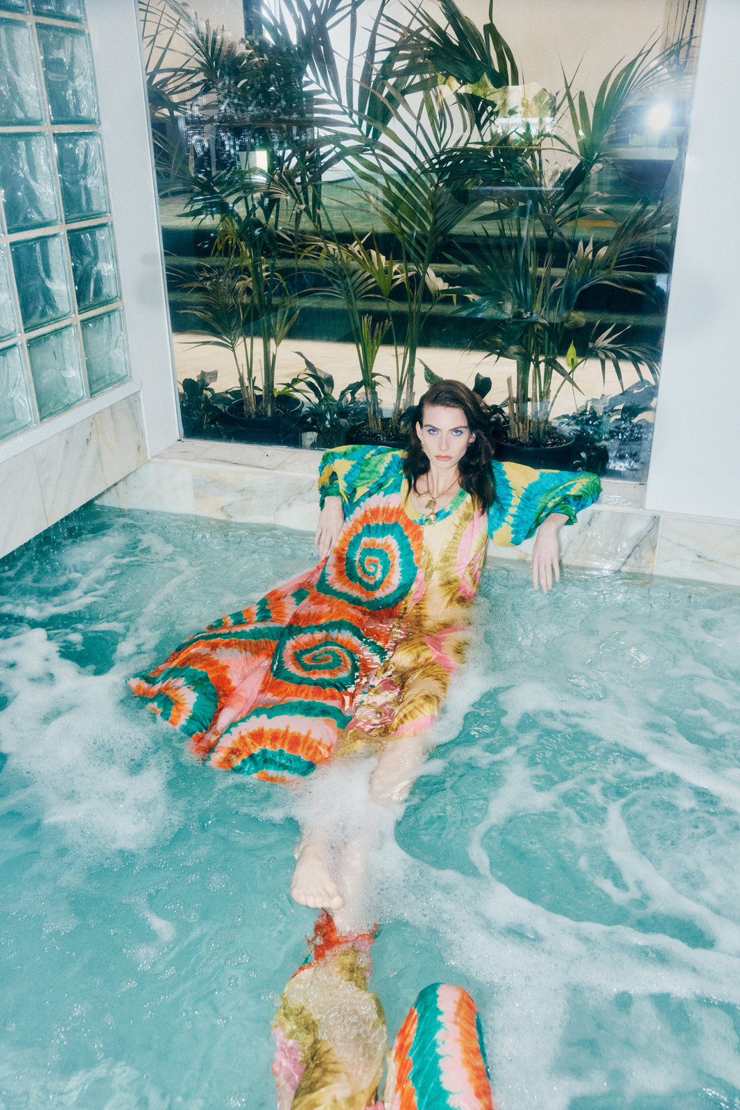 Woman in a vibrant, patterned dress sits in a pool with a reflective surface, palm trees, and textured tiles in the background.