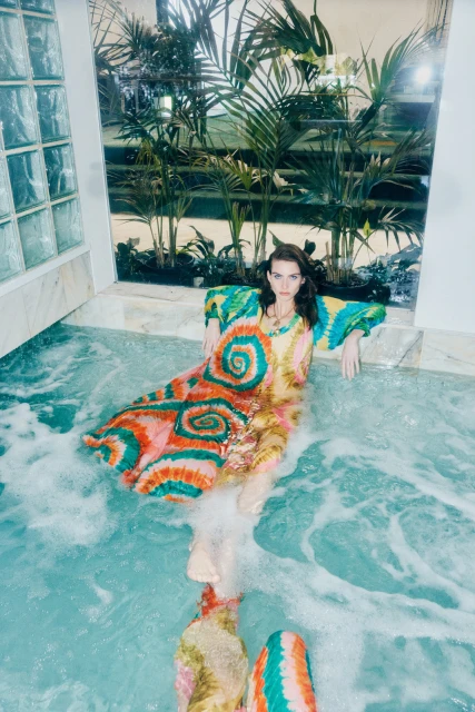 Woman in a vibrant, patterned dress sits in a pool with a reflective surface, palm trees, and textured tiles in the background.