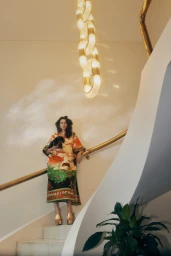 Model standing on a curved staircase holding a small dog, wearing a printed tunic and platform shoes under a modern chandelier.