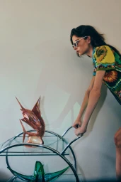 Woman wearing sunglasses and a vibrant floral dress leaning over a glass and chrome serving cart with glass sculptures.