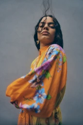 Low-angle shot of a woman in an orange floral dress with dark hair framing her face against a plain grey backdrop.