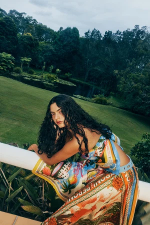 Woman with long dark wavy hair leaning on a white railing outdoors wearing a colourful patterned silk scarf top
