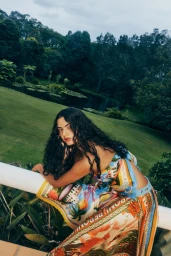 Woman with long dark wavy hair leaning on a white railing outdoors wearing a colourful patterned silk scarf top