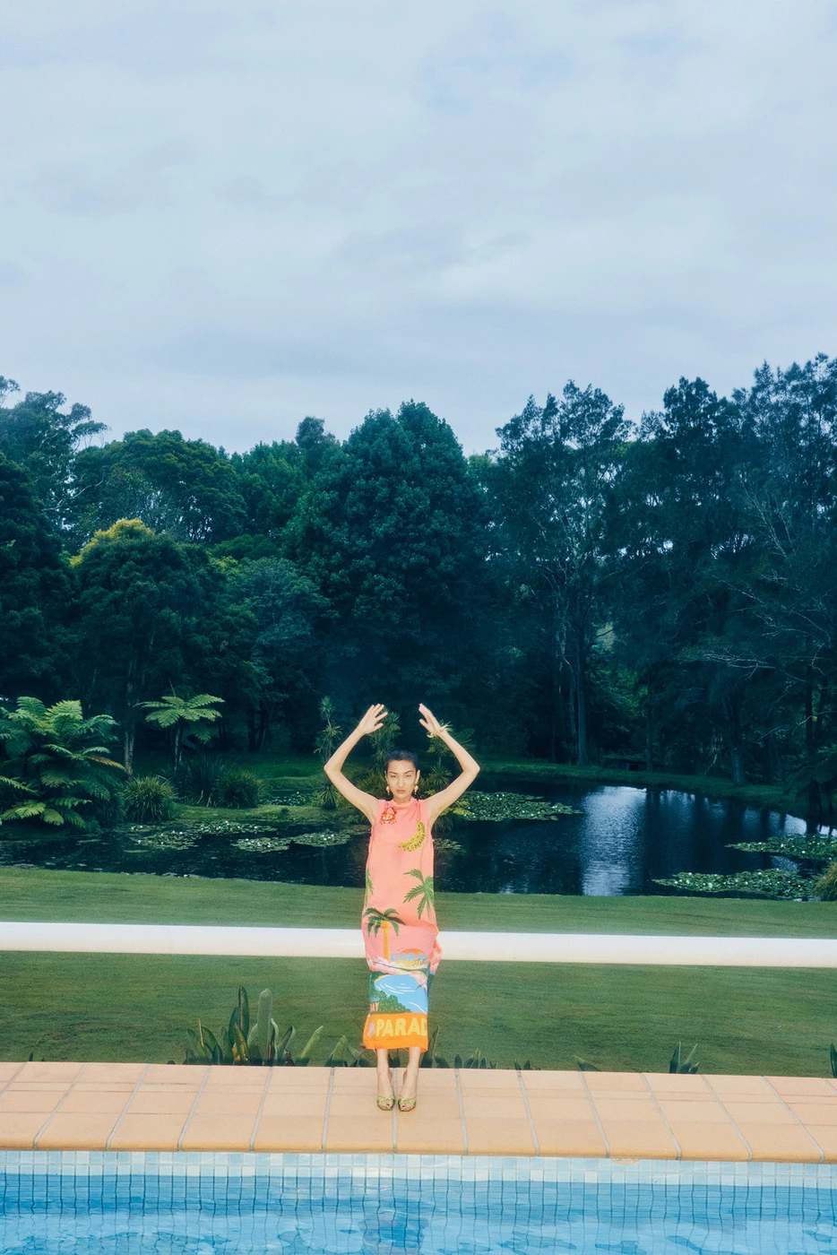 Model wearing a pink patterned dress standing by a swimming pool with a pond and lush trees in the background