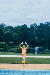 Model wearing a pink patterned dress standing by a swimming pool with a pond and lush trees in the background