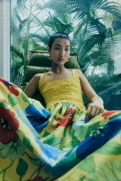 Model with short dark hair seated in a wicker chair wearing a yellow patterned floral dress against a tropical palm background.