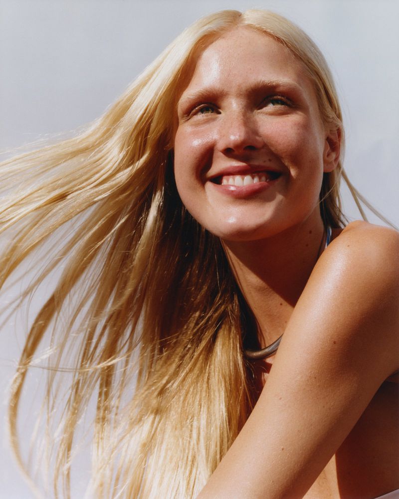 Close-up portrait of a smiling adult female with long blonde hair, wearing a necklace against a soft, bright background.