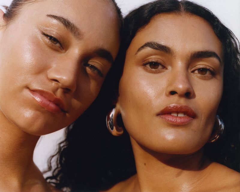 Close-up of two young adult women with neutral facial expressions. Their skin is glowing and bronze-toned. One woman has large hoop earrings.