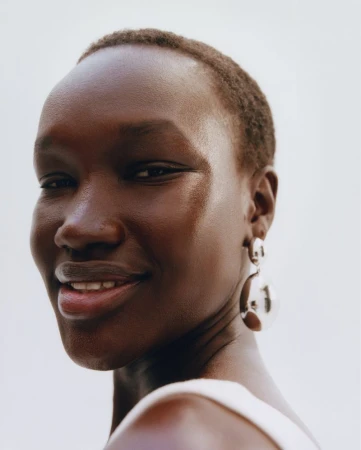 Close-up portrait of a smiling Black woman with short hair, wearing large silver hoop earrings and a white top against a white background.