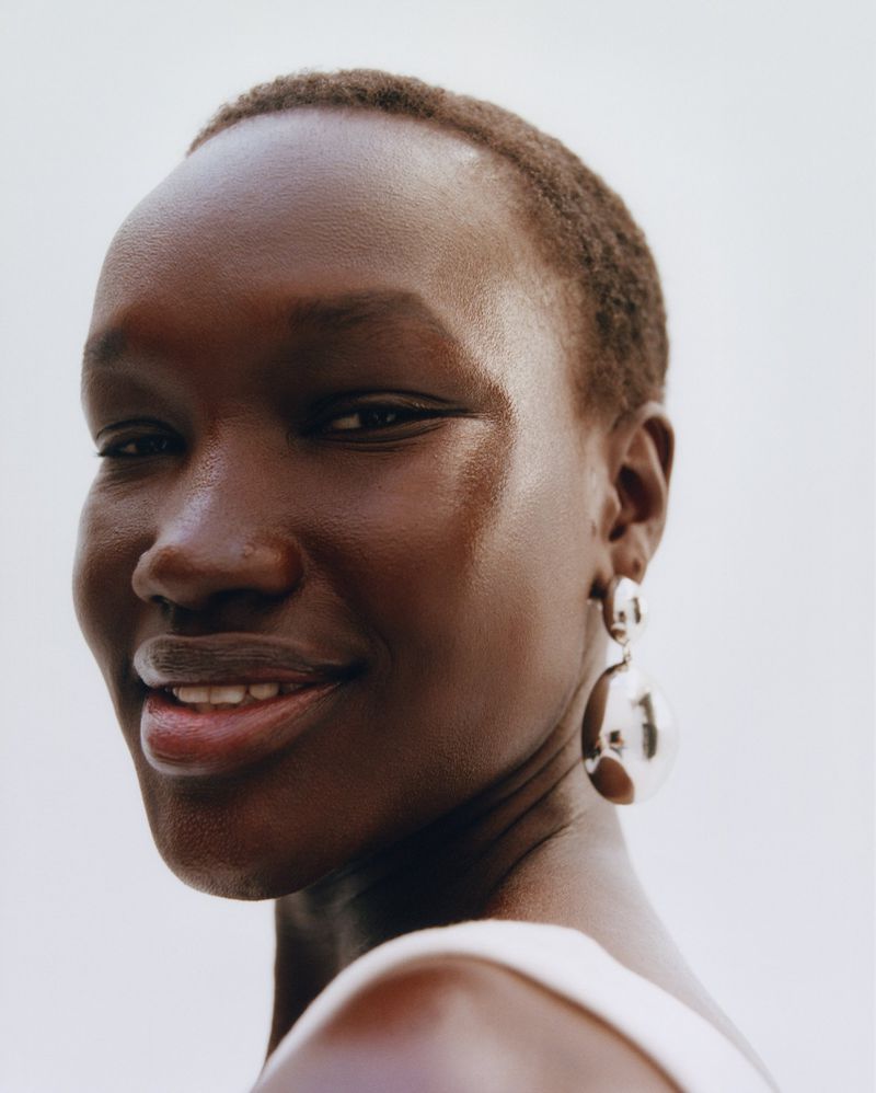 Close-up portrait of a Black woman smiling, showcasing her skin tone, short hair, and large silver earrings against a bright background.