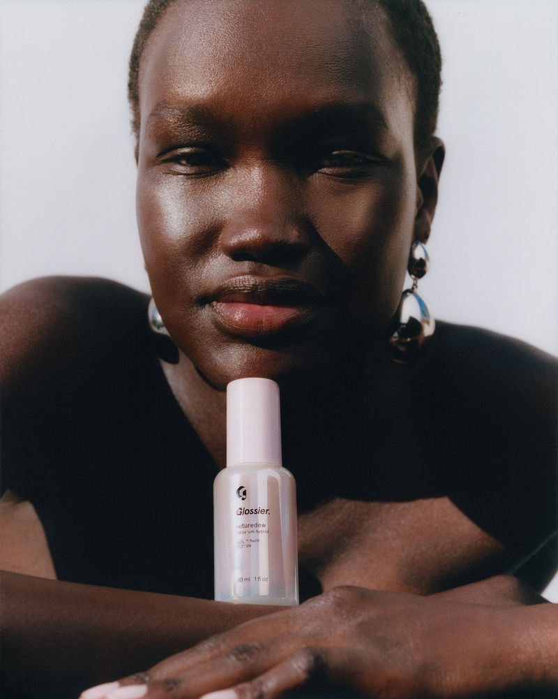 Adult female poses with a bottle of Glossier Rainbowfoil Haloscope in a beauty shot with studio lighting against a neutral background.