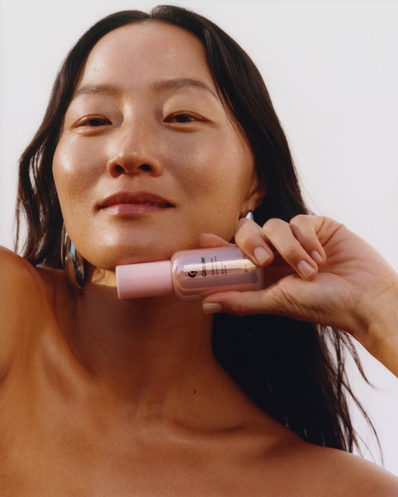 Woman holding a beauty serum bottle with light pink accents, against a plain white background in a beauty or skincare product shot.