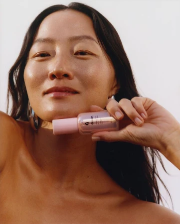 Woman holding a beauty serum bottle with light pink accents, against a plain white background in a beauty or skincare product shot.