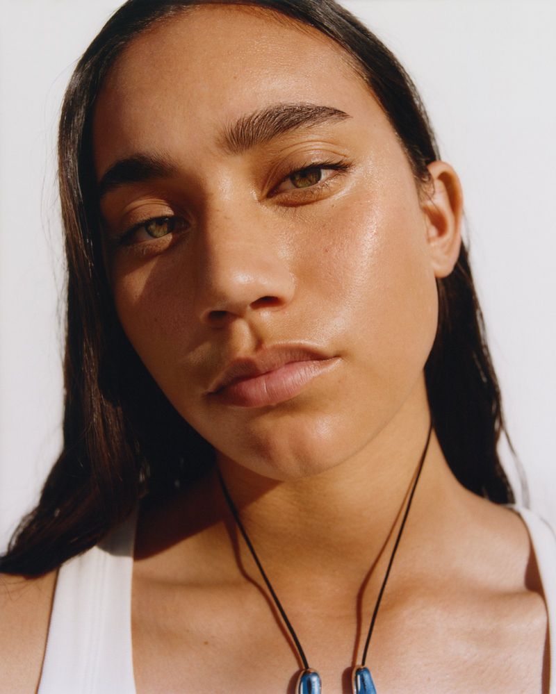 Close-up portrait of a young woman with long, dark hair and fair skin, wearing a white top and a blue pendant necklace.