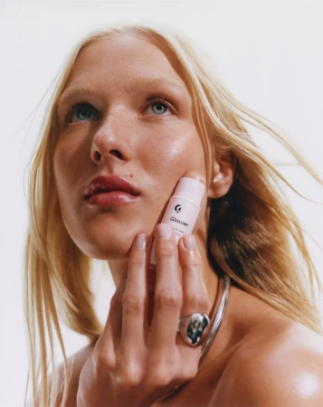 Close-up of a fair-skinned woman with blonde hair applying a pink cream stick to her cheek in a beauty advertising shot.