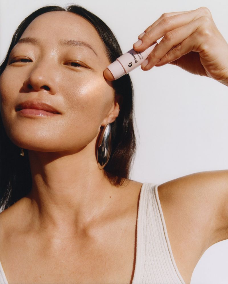 Portrait of a woman applying makeup stick on her cheek; she has large silver earrings and is wearing a light-colored top.