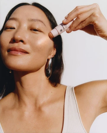 Portrait of a woman applying makeup stick on her cheek; she has large silver earrings and is wearing a light-colored top.