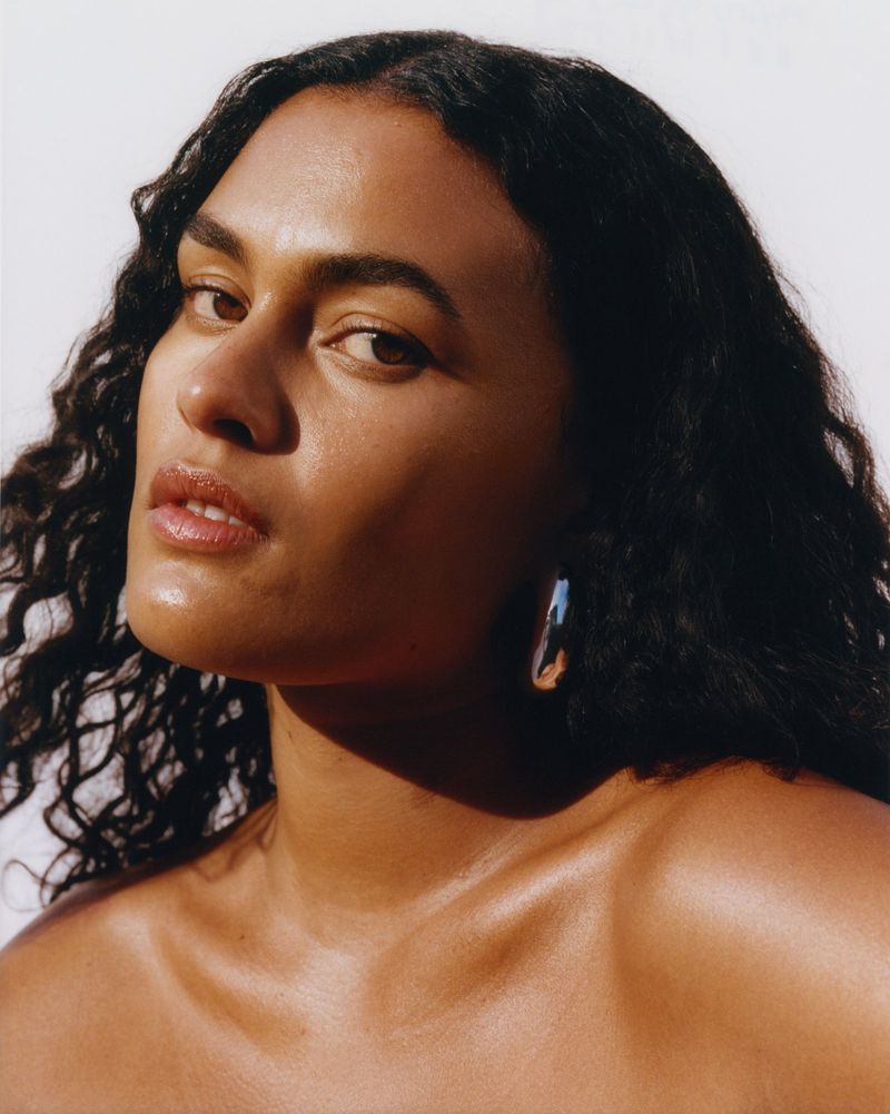 Close-up portrait of a brunette adult woman with dark, curly hair wearing hoop earrings in a photo studio setting with soft lighting.