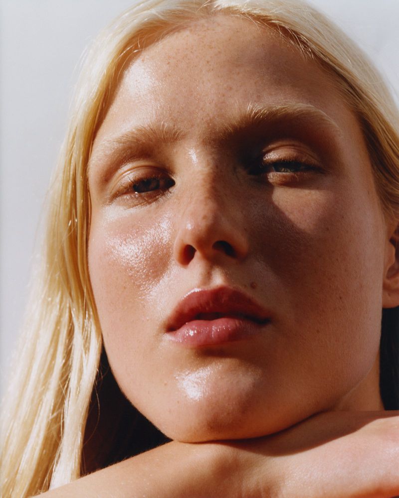 Close-up beauty portrait of a blonde woman with fair skin and freckles, gazing at the camera with natural light and slight shadows.