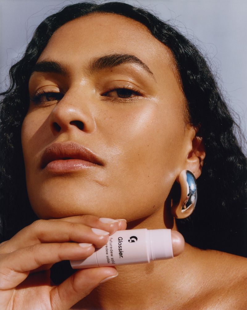 Close-up of a woman with dark curly hair holding a Glossier Haloscope highlighter stick, showcasing beauty and cosmetic product.