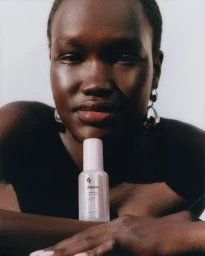 Model with dewy skin holding a Glossier Future Dew bottle against a white background