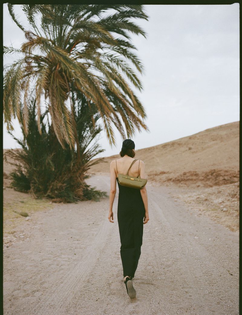 A woman in a black dress walks on a dirt path in a desert environment, with a palm tree and rolling hills under a cloudy sky.
