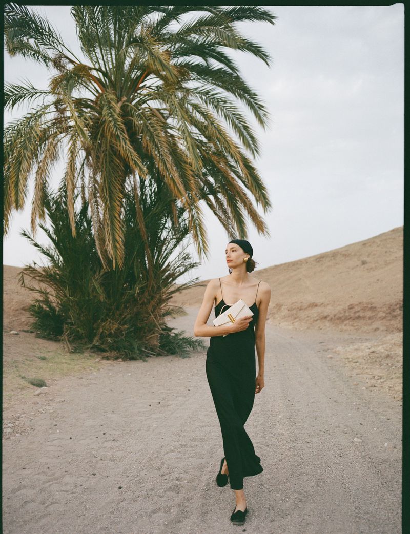 A full shot captures a woman with a clutch walking on a desert road, framed by a palm tree and sand dunes under an overcast sky.