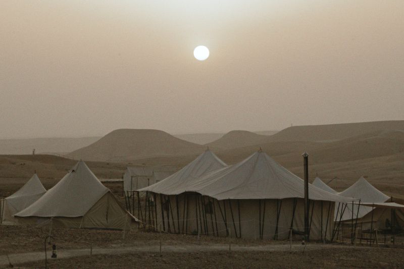Desert camping tents sit in a sandy landscape with a bright sun, creating a scenic and serene environment at sundown.