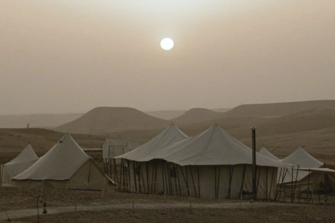 Desert camping tents sit in a sandy landscape with a bright sun, creating a scenic and serene environment at sundown.