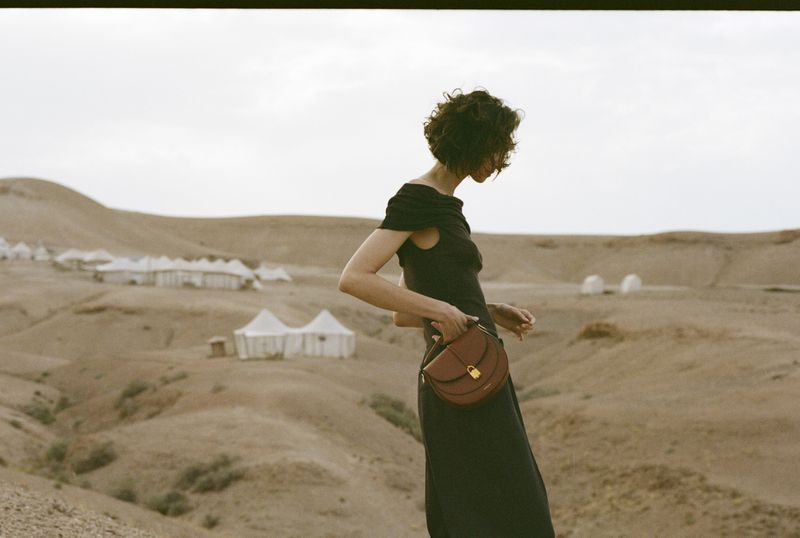 Adult female stands in desert landscape wearing an off-the-shoulder dress and carrying a brown handbag, with tents in the background.