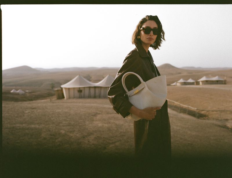 Woman in a trench coat and sunglasses holds a white leather bag outdoors against a background of desert tents and light sky.