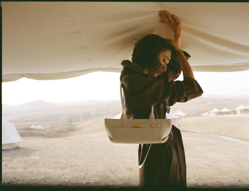 Adult female model in brown coat under canvas, holding white purse in desert landscape. Fashion editorial style outdoors.
