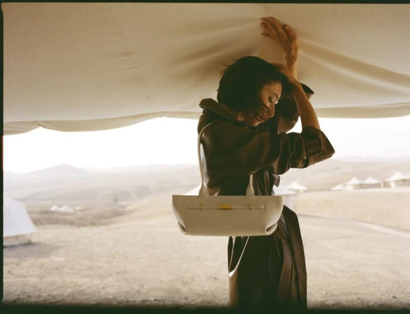 Adult female model in brown coat under canvas, holding white purse in desert landscape. Fashion editorial style outdoors.