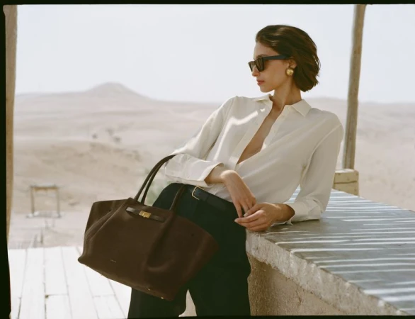 Model Alexandra Agoston in sunglasses holding a brown leather tote bag for DeMellier London S/S 2025 campaign.