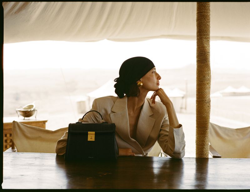 Woman in blazer with black purse sits at table in tent. Soft lighting highlights her face and jewelry in a desert-like setting.