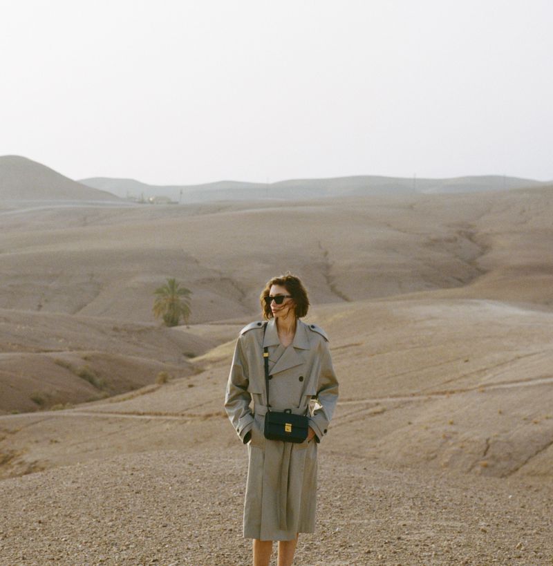 Woman in trench coat stands in desert landscape with palm tree in background, wearing sunglasses and crossbody bag on clear day.