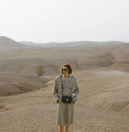 Woman in trench coat stands in desert landscape with palm tree in background, wearing sunglasses and crossbody bag on clear day.