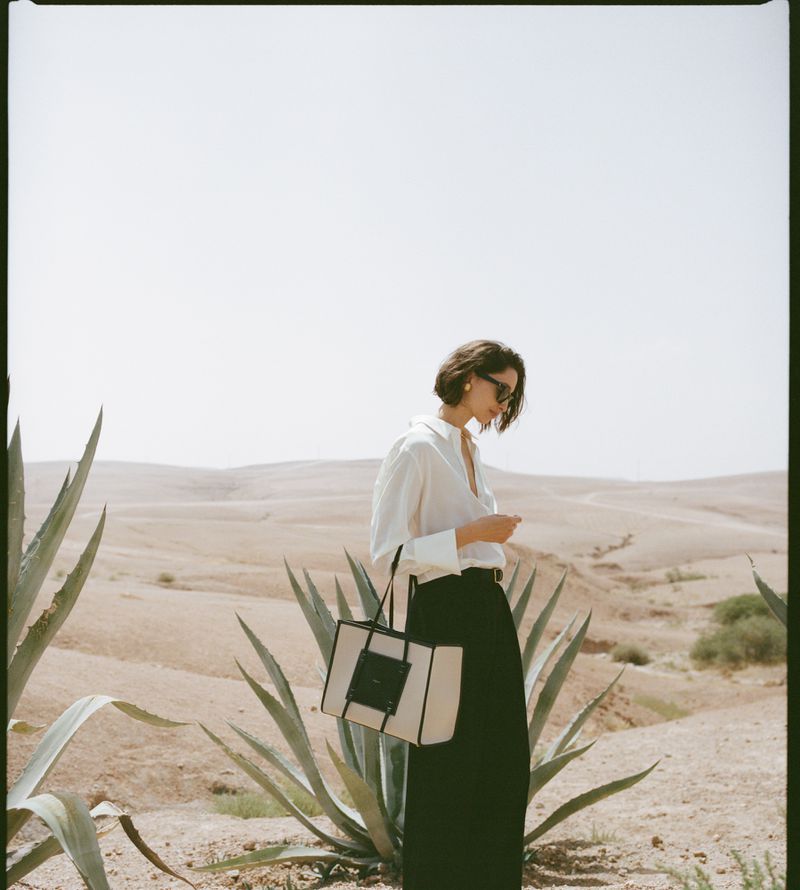 Adult woman in a white shirt and black pants stands outside in a desert landscape with a tote bag. She has short, dark hair.