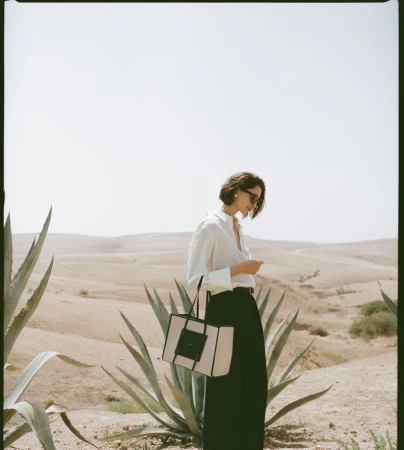 Adult woman in a white shirt and black pants stands outside in a desert landscape with a tote bag. She has short, dark hair.