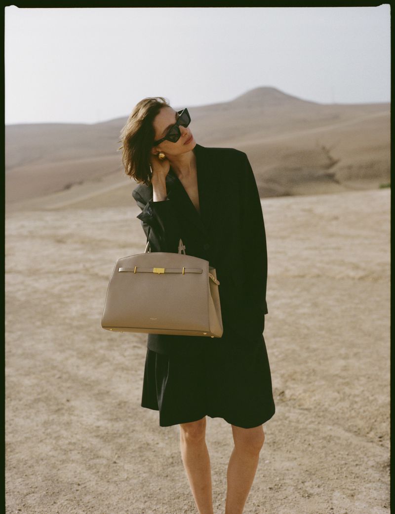 Model in black blazer wearing sunglasses holding a beige handbag against a desert landscape