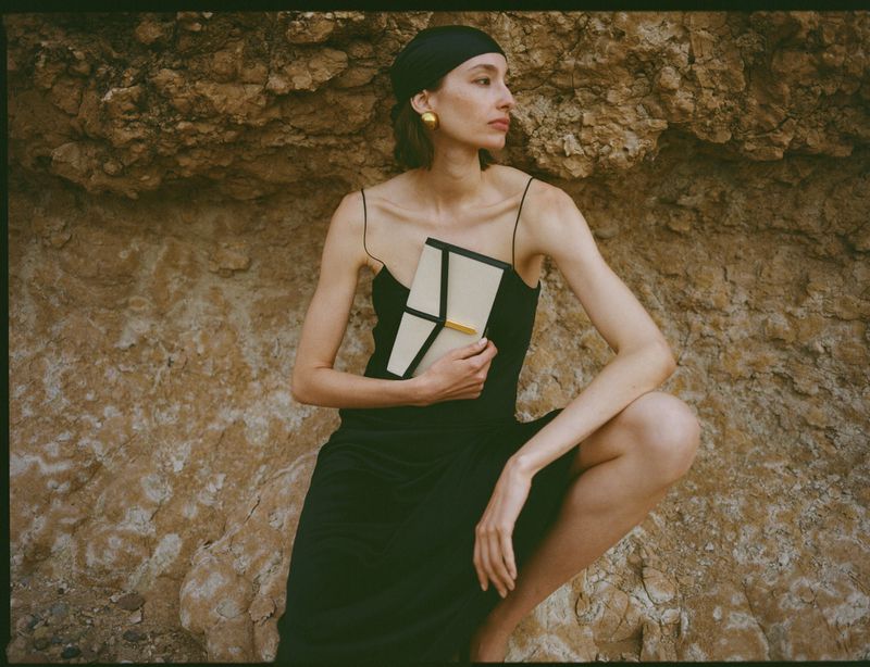 Model wearing a black dress and headscarf holding a structured handbag against a rough rock cliff background.