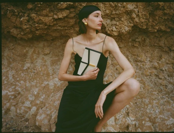 Model wearing a black dress and headscarf holding a structured handbag against a rough rock cliff background.