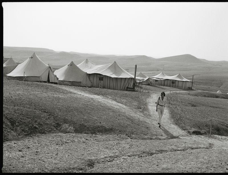 Woman walking on a desert path near a tent camp. Landscape includes tents, dirt path, arid ground, and distant mountains in monochrome.
