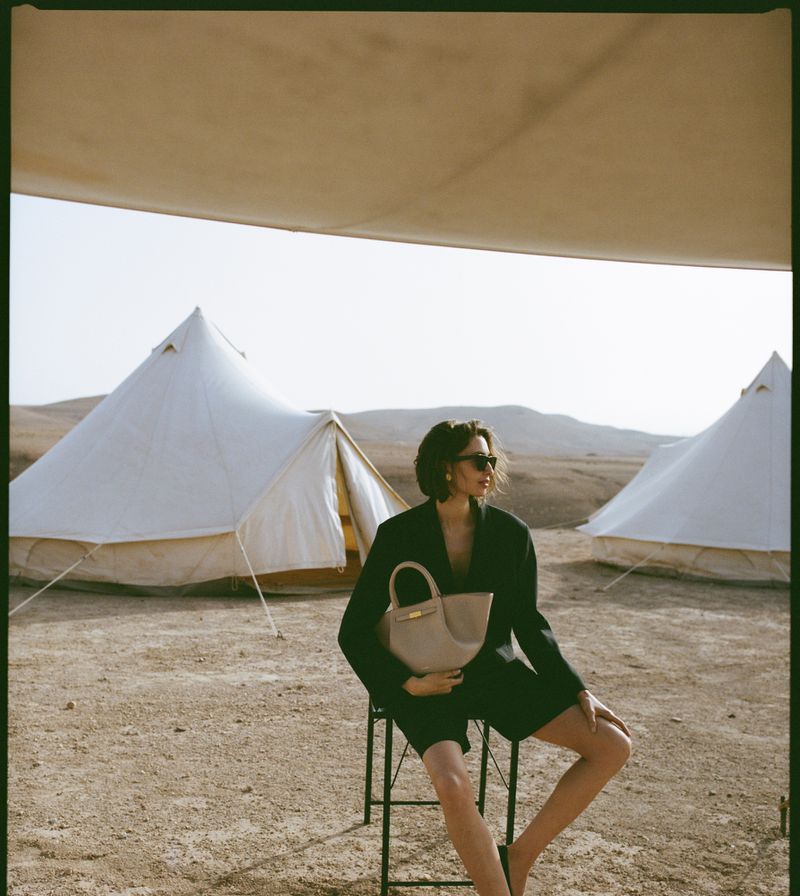 A woman sits on a stool in the desert with white tents in the background, wearing a black blazer, shorts, and holding a beige bag.