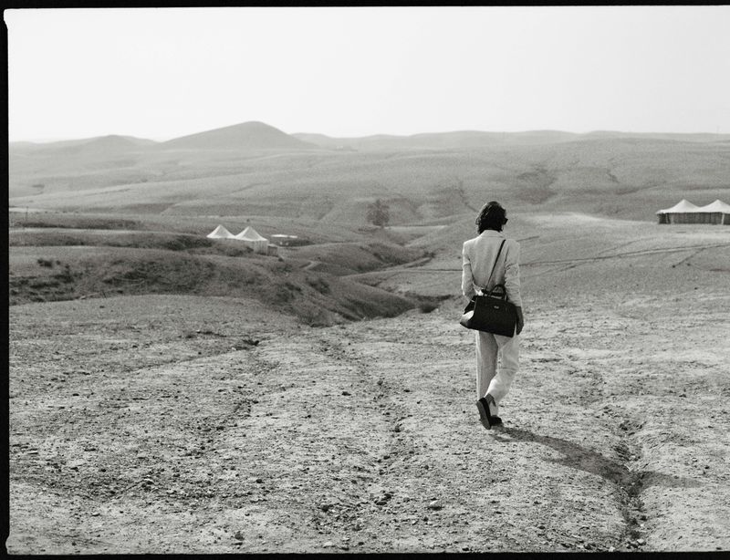 Person walks across arid landscape towards tents. Wearing light suit and carrying bag, monochromatic scenery, hills, desert.