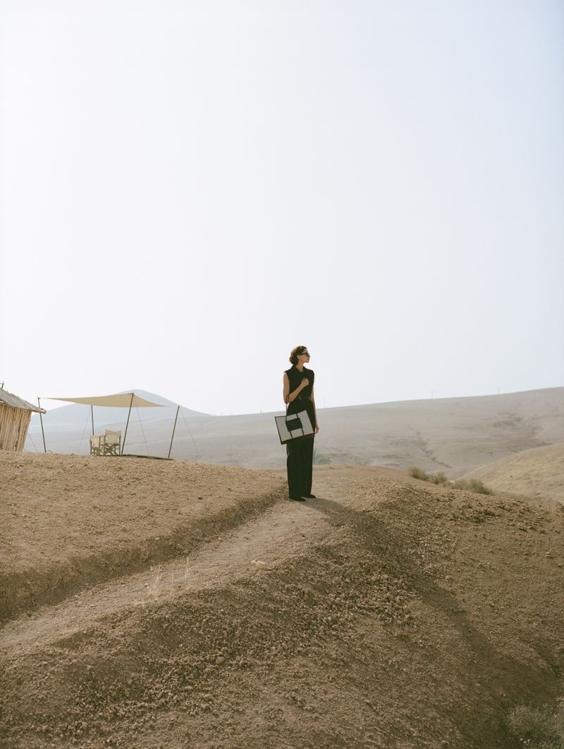 A woman in a black dress stands on a sandy hill holding an abstract art piece in a desolate landscape under a bright, clear sky.