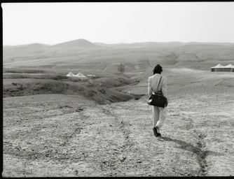 Model walking away across a vast desert landscape carrying a dark shoulder handbag for DeMellier London SS 2025 campaign.