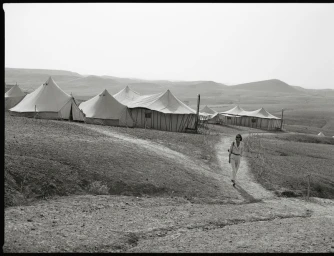 Model walking along a dirt path past a camp of large tents in a sparse landscape for DeMellier London S/S 2025 campaign