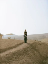 Model wearing dark clothing and holding a handbag on a desert hill under a bright sky, photographed by Sonia Szóstak for DeMellier London.