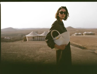 Model Alexandra Agoston wearing sunglasses and a leather coat holds a white handbag in a desert landscape for DeMellier London.
